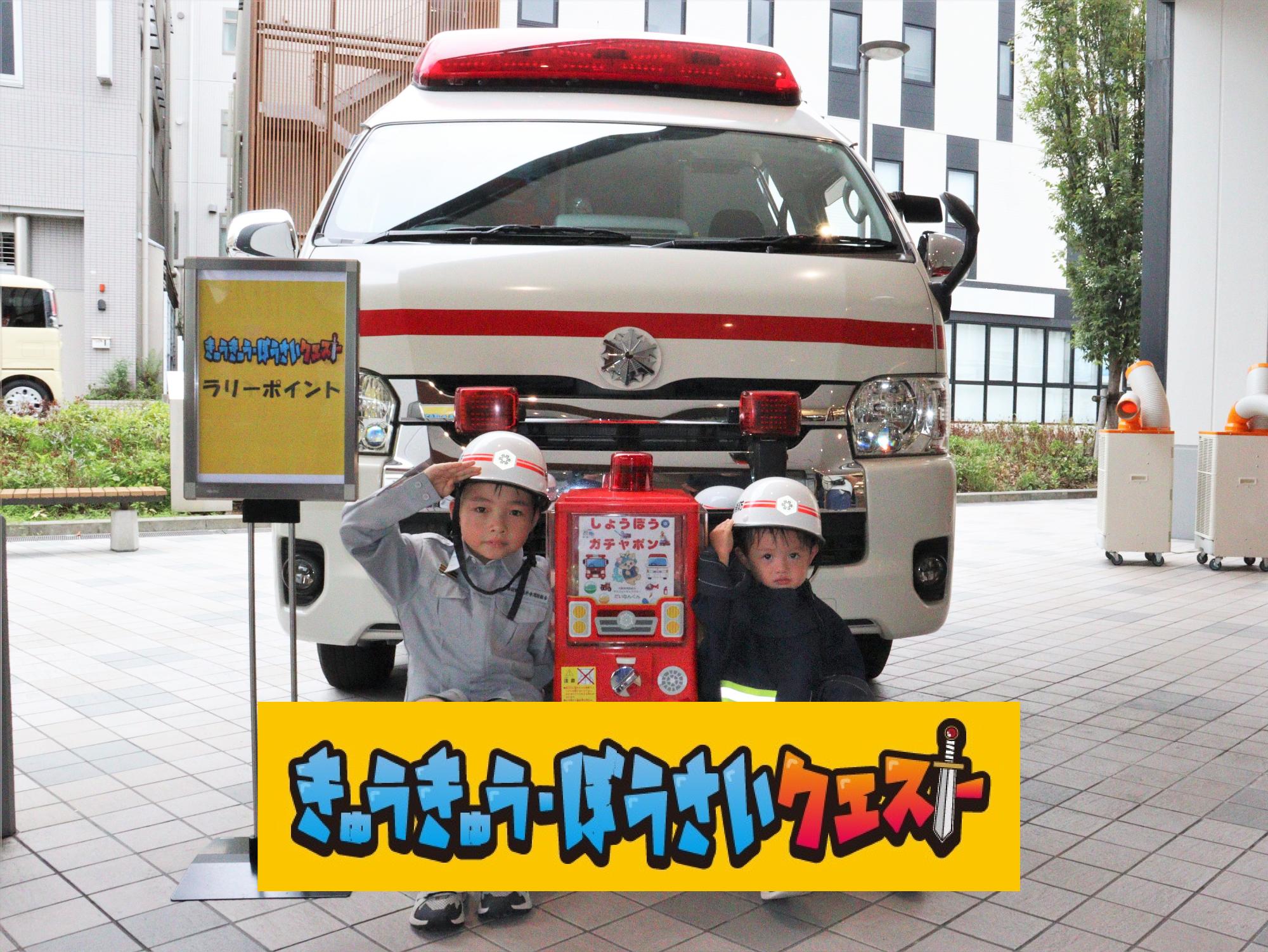 救急車の前で消防服に着替えた子供２人が記念撮影している様子