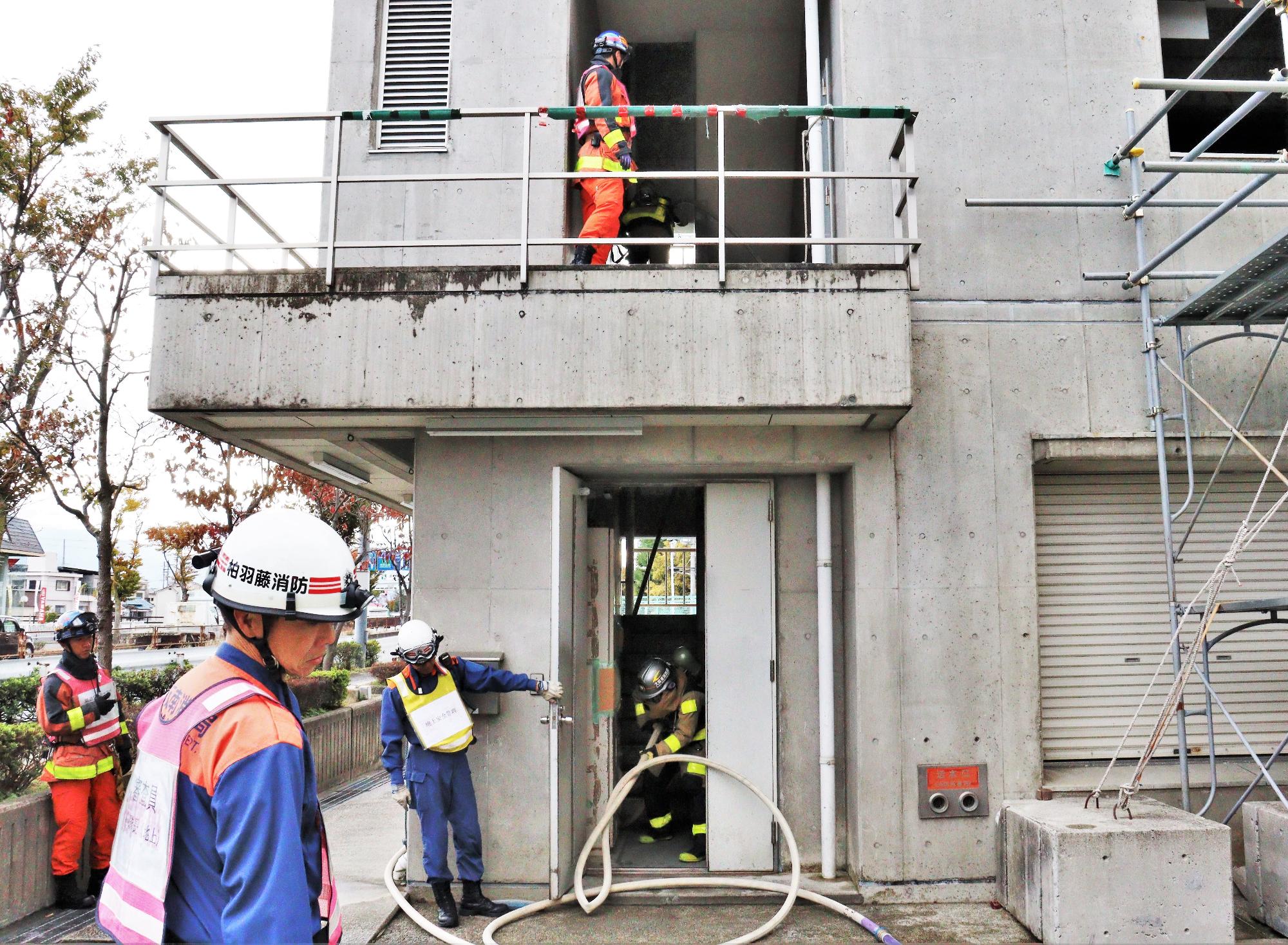 隊長と隊員1名が火災建物の階段をホースを携えて駆け上がりもう一人の隊員が玄関から消防ホースを引っ張っている様子